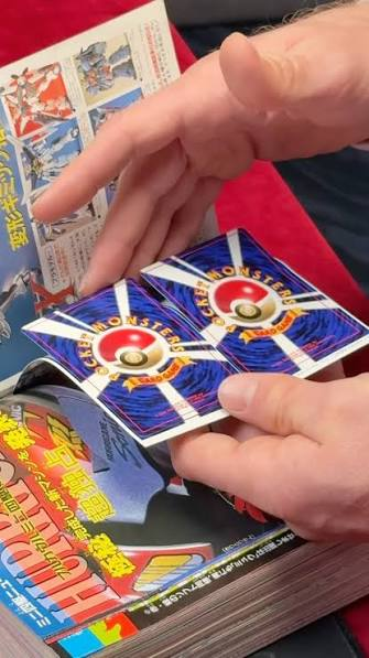 Two collectible trading cards featuring a colorful design with a Poké Ball icon, held in hands above a stack of magazines.
